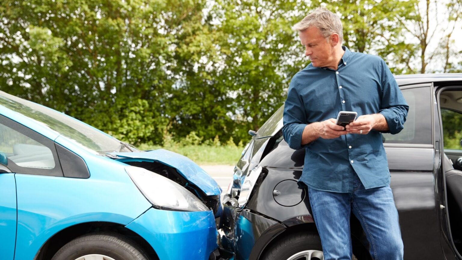 Male driver standing by damaged cars after accident, using phone to report insurance in car accident claims.
