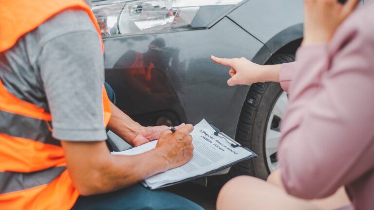 Insurance agent filling out a vehicle damage claim form while driver points to damage
