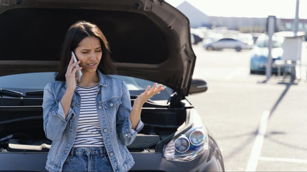 Young woman standing by a car with the hood open while making a phone call